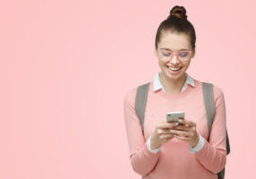 Close-up portrait of young female student looking excited at display of smartphone, smiling and laughing, impressed by social media content, isolated on pink background